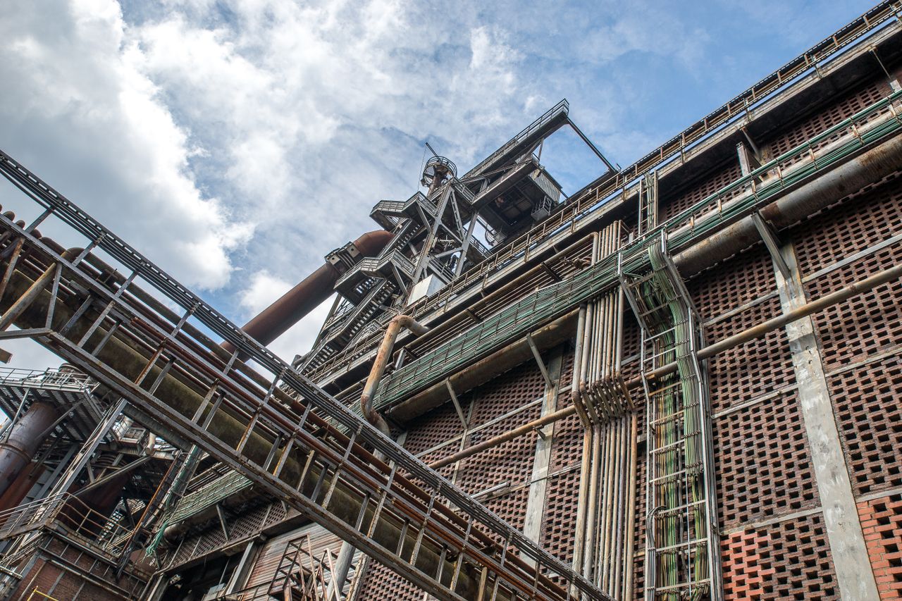 A detailed view of a metal industrial structure with pipes and beams under a cloudy blue sky.